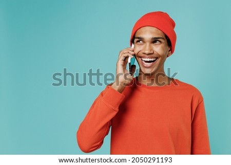 Young friendly smiling happy african american man in orange shirt hat talk speak on mobile cell phone conducting pleasant conversation isolated on plain pastel light blue background studio portrait.