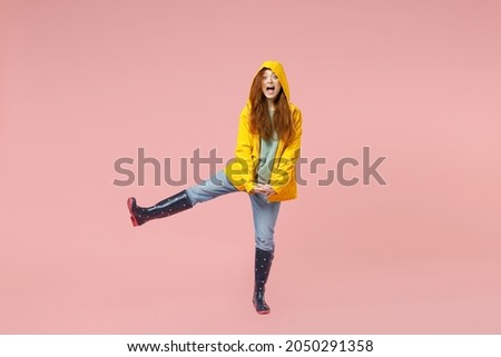 Full length redhead young woman in yellow waterproof raincoat hood outerwear outstretched leg fingers intertwined isolated on pastel pink background Outdoor lifestyle wet fall weather season concept