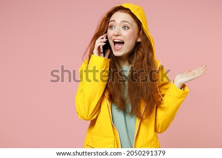 Redhead young woman in yellow waterproof raincoat hood outerwear talk speak by mobile cell phone spread hand isolated on pastel pink background studio Outdoor lifestyle wet fall weather season concept