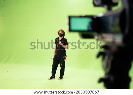 Selective focus of Asian male studio staff wear a face mask and walkie-talkie at the pants, holding a paper roll standing on a green screen background with a blurred video camera in the foreground.