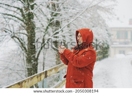 Middle age man holding smartphone, taking pictures or video for social media, winter weather with snowfall