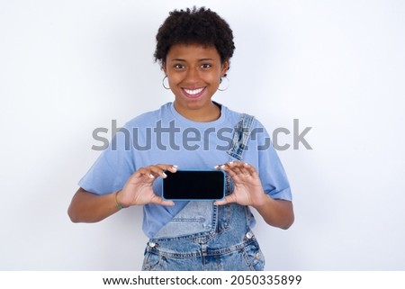 Cheerful cheery content African American woman with afro hair style wearing denim overall against white wall holding in hands device hobby smm post blog