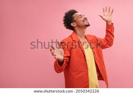 Medium shot of happy African young and smiling man with closed eyes with copy space. Well dressed in fall colors with arms raised at his sides against solid pink background. Royalty-Free Stock Photo #2050342226