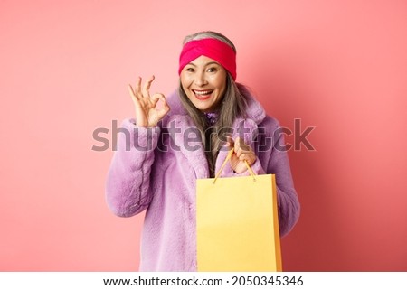 Shopping and fashion. Happy asian senior woman in stylish clothes holding paper bag from store, showing OK sign, recommending shop, pink background