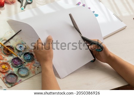 cutting a blank paper with scissors on a desk with working tools for crafts such as palette with colors and brushes, art and creativity instead of work, lifestyle