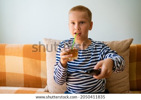 Young man watches tv, using remote control to change channels. Guy got bored with what he sees on tv screen. Sitting on the couch in your living room at home, copy space. Drinks juice from a glass