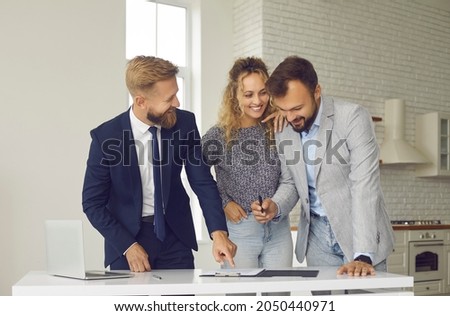 Happy young married couple buying or renting new home. Smiling husband and wife standing at table in kitchen of their new house or apartment and signing contract given by realtor or real estate agent