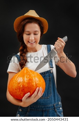 Woman in overalls on a black background cuts a lantern out of a pumpkin with a knife for the Halloween holiday.