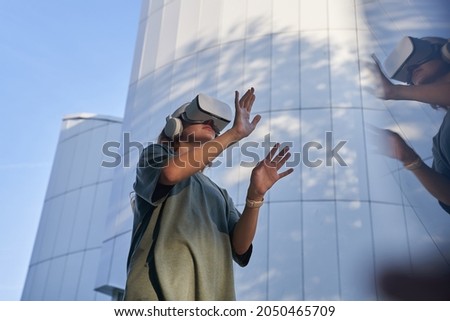 A girl is having fun in virtual reality glasses, standing against the background of a mirrored building. He meets his hands in the reflection. The concept of the future. High-quality photo