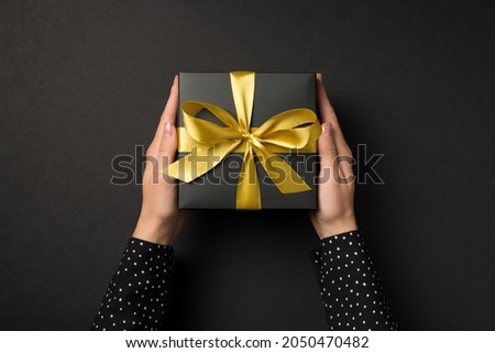 First person top view photo of hands in polka dot shirt holding black giftbox with golden satin ribbon bow on isolated black background Royalty-Free Stock Photo #2050470482