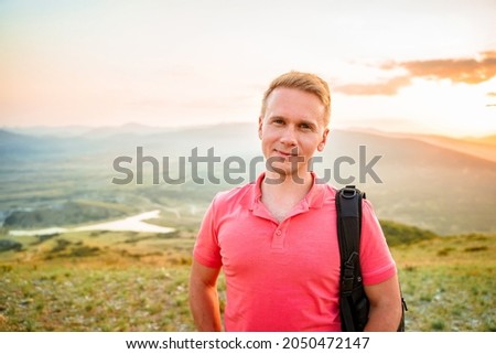A young blond man in a pink shirt conquered the top of the mountain at sunset. Beautiful mountain landscape with a gradient. The concept of enjoying nature.