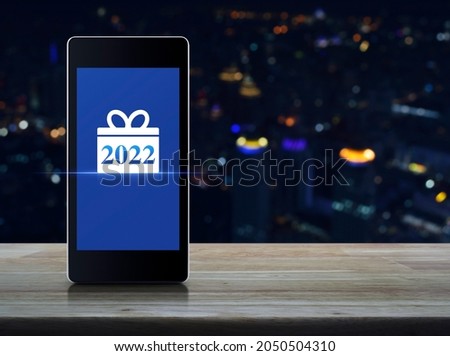 Gift box happy new year 2022 flat icon on modern smart mobile phone screen on wooden table over blur colorful night light city tower and skyscraper, Business happy new year 2022 shop online concept
