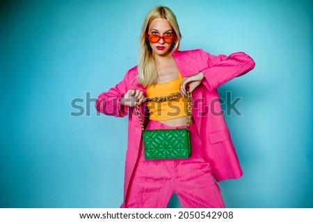 Fashion portrait of confident woman wearing trendy summer pink fuchsia color suit, orange sunglasses, holding green quilted faux leather bag, posing in studio, on blue background. Copy, empty space
 Royalty-Free Stock Photo #2050542908