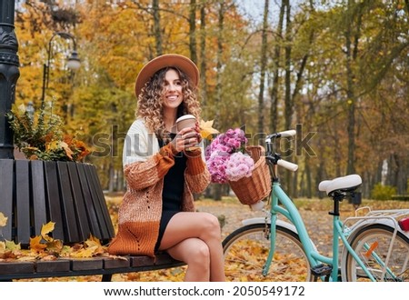 Morning coffee break in city park with bike at autumn. Likable young lady sitting on round bench outdoors with her women's city bicycle admiring autumn landscape. Royalty-Free Stock Photo #2050549172