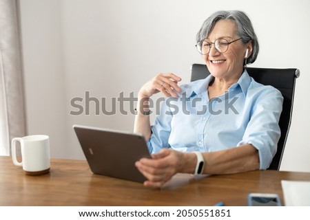 Attentive elder female office worker businesswoman in glasses sitting at the desk, browsing the internet, listening to a podcast, checking emails from the clients, using discovering new corporate app