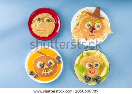 Creative homemade Halloween pancakes for breakfast, in form of funny monsters, ghost, bat, witch. With traditional trick or treat sweets, candy and decorations, top view on colorful blue background