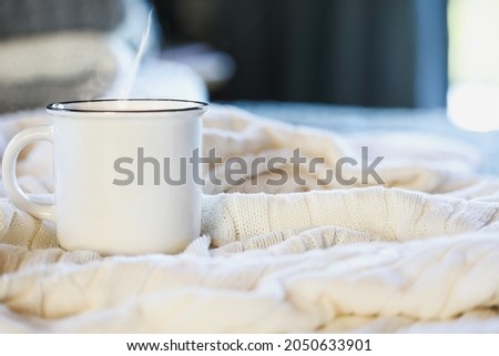 Hot steaming cup of coffee sitting on top a soft white knit blanket on a bed with stack of covers in background. Selective focus with extreme blurred foreground and background. 