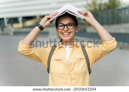 Education concept. Funny caucasian student girl laughing out loud and posing with book on her head while standing at the street. Stock photo