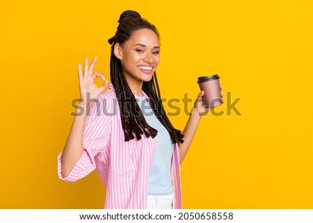 Photo of young happy positive dark skin woman make okay sign hold coffee cup wear striped shirt isolated on yellow color background