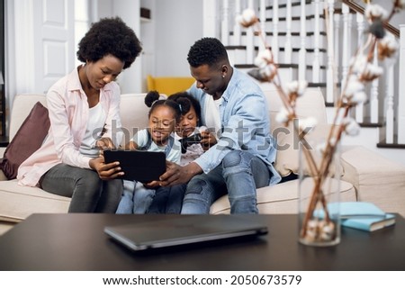 African parents and their two cute daughters watching cartoons on digital tablet while resting together on comfy couch. Concept of family, technology and leisure time.
