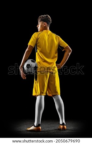 Back view full-length portrait of young handsome football player in yellow uniform isolated over black background. Concept of action, team sport game, energy, vitality. Copy space for ad. Royalty-Free Stock Photo #2050679690