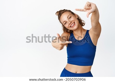 Smiling fitness woman in activewear, looking through hand frames as if taking photo, picturing moment, standing in workout clothing, white background