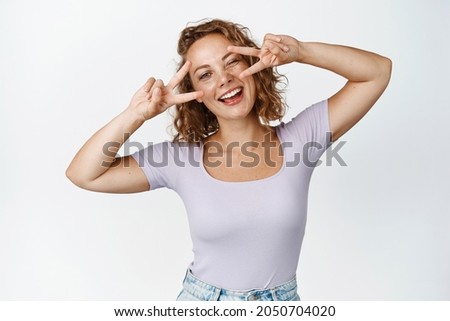 Beautiful young natural woman with curly blond hair, winking, showing v-sign peace near eyes, smiling happy, white background