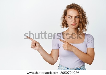 Disappointed young woman with short curly hair, frowning and sulking, pointing fingers left with dislike face, white background