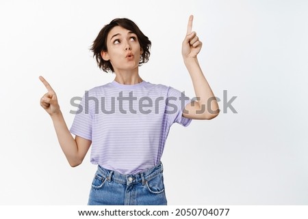 Curious girl seeing smth interesting, pointing sideways but look up way to product, say wow, picking product in shop, making her choice, standing over white background
