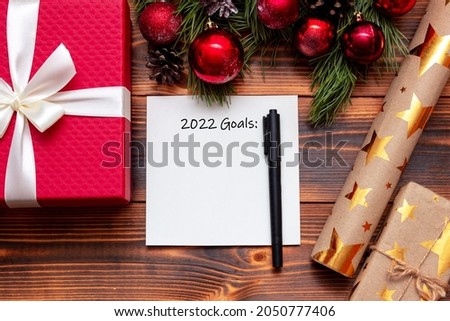 A white sheet with the title 2022 goals, a black marker on a wooden table with Christmas, New Year's decorations: gift boxes, a craft wrapper with gold stars, a branch of a fir tree with red toy balls