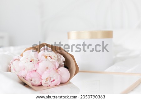 Gold gift box and white frame mockup on the bed. Bouquet of pink peonies in craft packaging. Scandivanavian white interior