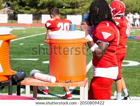 An African American high school football player drinking water out of a cup next to an orange cooler on the sidelines during a football game. Royalty-Free Stock Photo #2050809422