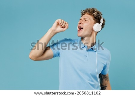 Jubilant young curly man 20s years old wears azure t-shirt listen music in headphones dance sing song in hand like in microphone have fun isolated on plain pastel light blue background studio portrait