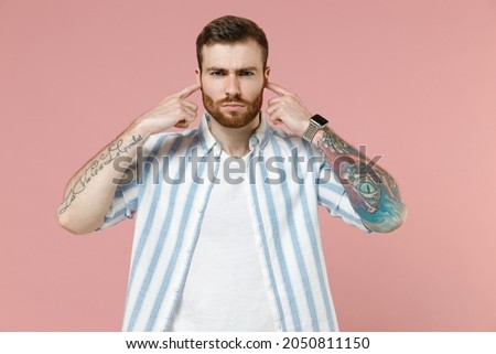 Young sad caucasian unshaven man 20s in blue striped shirt white t-shirt cover ears with hands fingers do not want to listen isolated on pastel pink background studio portrait Tattoo translate fun.