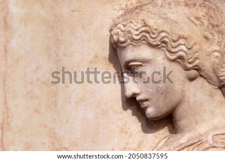 Ancient Greek relief of woman on marble wall with copy space for text. Remains of culture of past civilization in Greece. Beautiful stone sculpture, side view of face, old Greek art background. Royalty-Free Stock Photo #2050837595