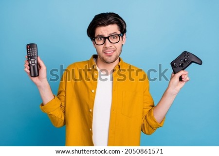 Photo of uncertain puzzled guy hold remote control gamepad make choice wear yellow shirt isolated blue color background
