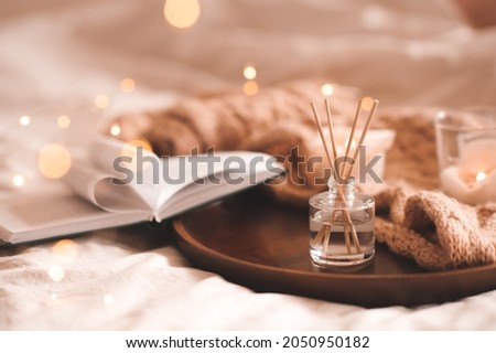 Home perfume in glass bottle with wood sticks, scented burn candles, open paper book and knit wool textile on ray in bedroom close up. Aromatherapy cozy atmosphere lifestyle. Winter warm xmas season.  Royalty-Free Stock Photo #2050950182