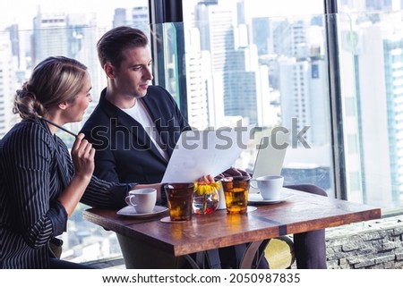 Picture of young business man and woman taking to each othe. They are in white shirt and suit. They are in a hotel lobby. 