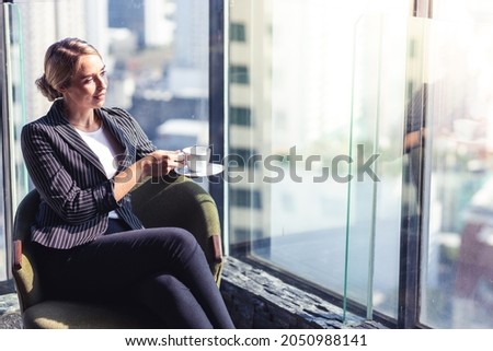Picture of young business woman holding a cup of coffee. She is wearing white shirt and suit. She is sitting in a hotel lobby.