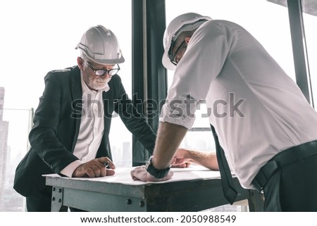 Picture of two engineer discussing their construction project together in a hotel lobby. They are wearing white shirt , suit and hardhat. They are drawing on a piece of paper with pen in their hands.
