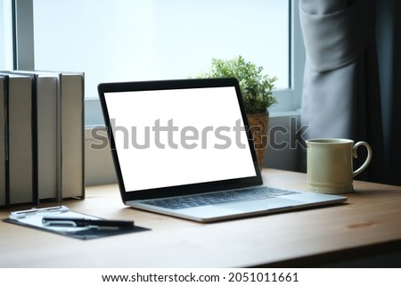 Mock up of white blank screen laptop putting on a wooden table surrounded by a coffee cup, clipboard, pen, potted plant and books with the window and curtains as a background.