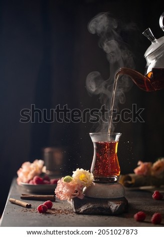 Hot berries tea pouring from the teapot into the cup on the table with fresh raspberries,cinnamon sticks and beautiful gentle flowers.Close up of hot drink in freeze motion.