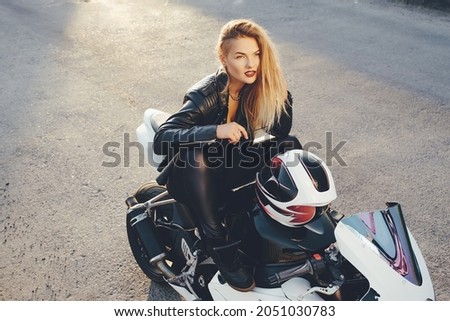 Rebel freedom concept. Young beautiful biker woman sitting with bent legs on a motorbike in a fashionable pose. Woman wearing leather clothes sitting on a white motorcycle holding arm on helmet