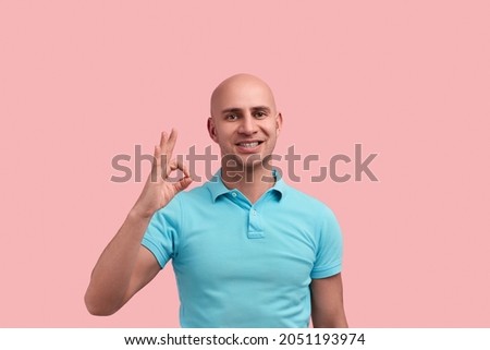 Cheerful happy bald homosexual man with bristle shows ok gesture, says okay, raises hand, smiles, recommends product, looks straight, gay friendly, wears blue polo shirt, stands over pink background