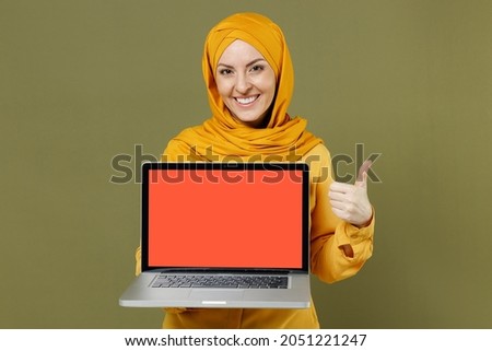 Young arabian asian muslim woman in abaya hijab yellow clothes hold laptop pc computer blank screen workspace show like gesture isolated on olive green background People uae islam religious concept