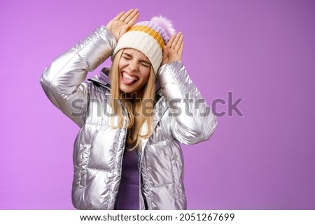 Carefree funny amused fair-haired caucasian girl 25s having fun playing around mimicking bunny close eyes show tongue excited make animal ears palms on head standing purple background joyful