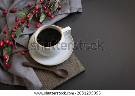 Coffee mug filled with black coffee on saucer and cutting board with red berries and copy space on black background 