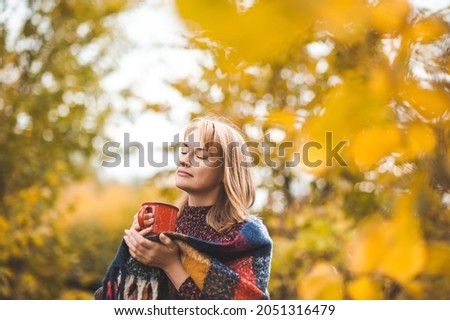 A beautiful blonde woman drinks a hot drink from a red mug in an autumn park. The woman is wrapped in a warm plaid scarf. Royalty-Free Stock Photo #2051316479
