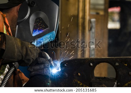 Man dressed in orange coveralls working with an arc welder on a piece of steel held in a vise