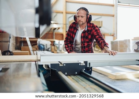 Carpenter in a woodworking workshop or factory using large industrial machinery during the production process wearing ear muffs against noise Royalty-Free Stock Photo #2051329568